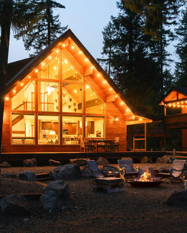 Glowing A-frame cabin at dusk with fire pit and string lights in the pines