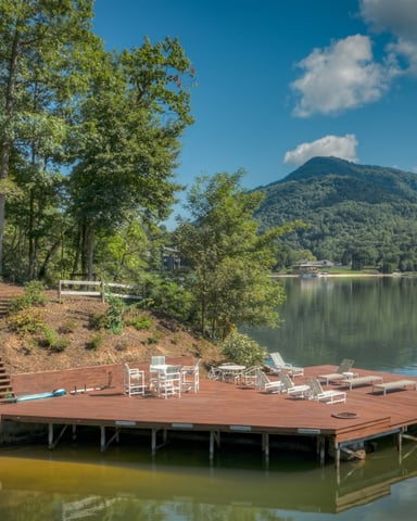 Waterfront deck with lounge chairs and dining furniture overlooking calm lake, forested mountain in background