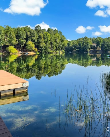 Wooden dock extending into calm lake with forested shoreline and blue sky reflected in water