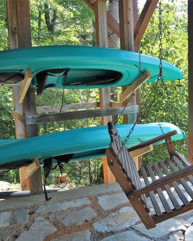 Two turquoise kayaks suspended from wooden pergola beams above stone patio, surrounded by green trees
