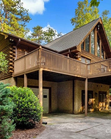 Modern log cabin with peaked roof, wraparound deck, and stone foundation surrounded by tall trees