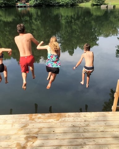 Four children running into calm river water from wooden dock, surrounded by green trees and grassy banks.