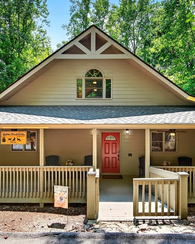 Tan cabin with red door and wraparound porch surrounded by dense green forest