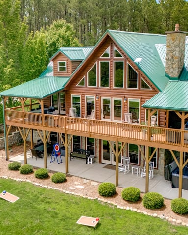 Large log cabin with turquoise metal roof, wraparound deck, stone chimney, surrounded by forest and manicured lawn.