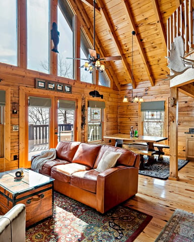 Spacious log cabin interior with vaulted wood ceiling, leather seating, large windows, and open kitchen area