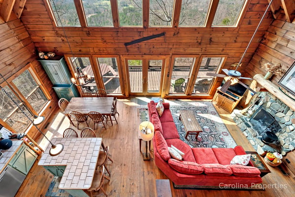 Overhead view of wood cabin interior with red sofa, dining table, kitchen, and expansive windows overlooking forested mountains