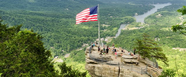 American flag flying on rocky cliff overlook with river valley and forested mountains below
