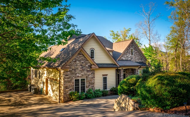 Stone and siding house with peaked roof and attached garage surrounded by trees and landscaping